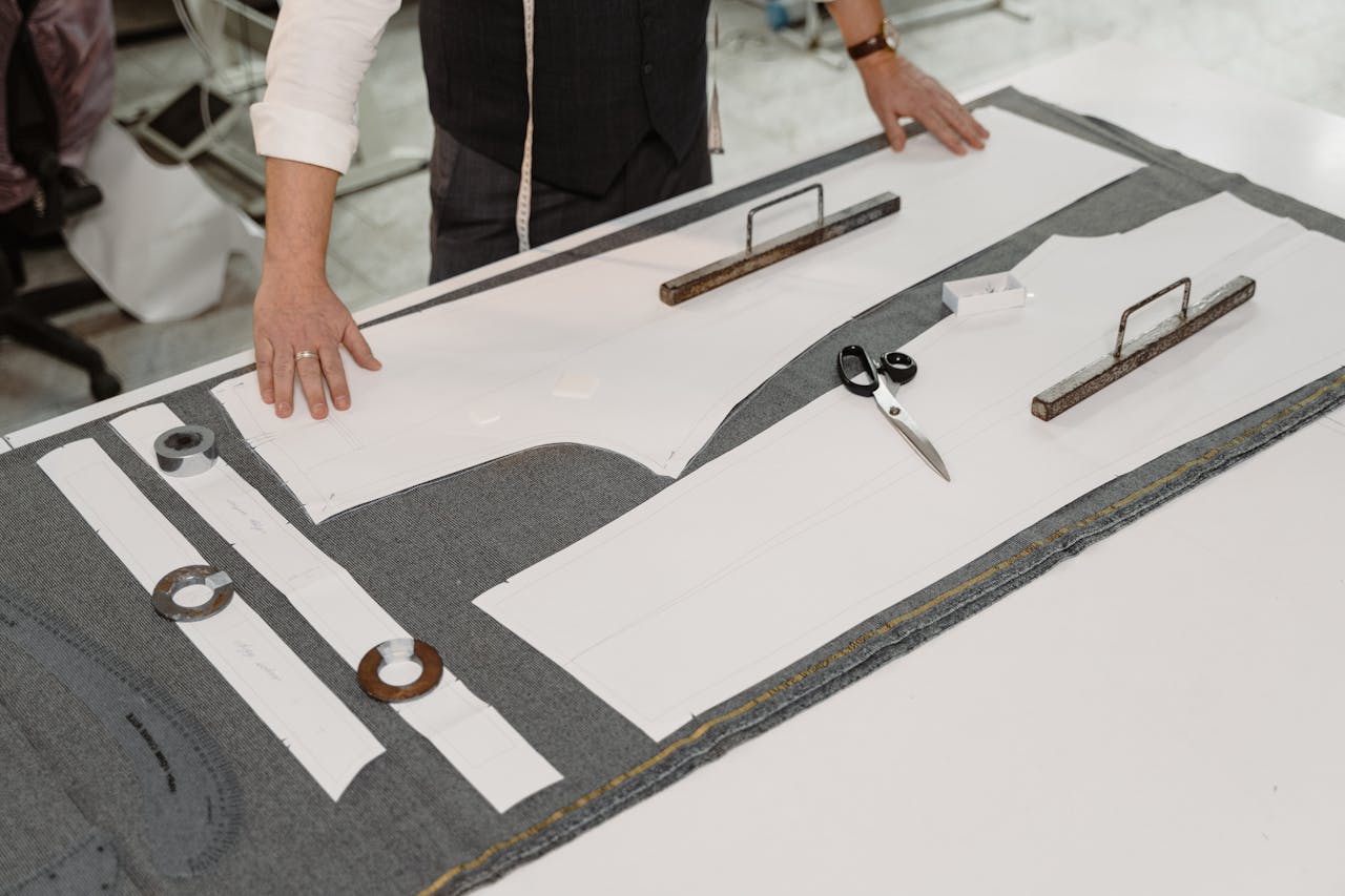 heros-img Hands of a tailor working with fabric and patterns in a workshop, showcasing traditional craftsmanship.